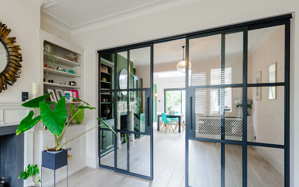 Period living room opening through steel-framed glass doors to a bright dining room, with built-in shelving and a large-leaf plant